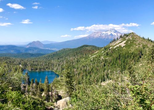 A scenic view of a mountain landscape featuring a lake, lush forests, and distant peaks under a clear blue sky.