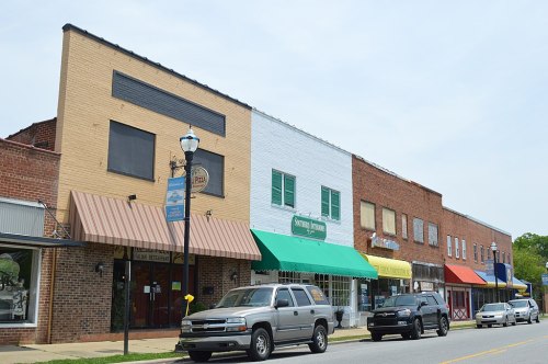 A row of colorful storefronts along a street, with parked cars and a lamppost in the foreground.