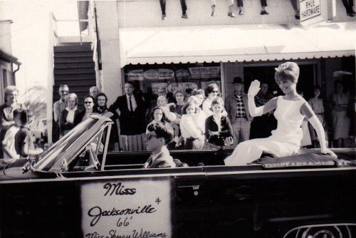 A vintage parade scene featuring a woman in a convertible, waving to a crowd, with a sign reading "Miss Jacksonville."