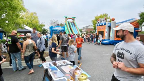A lively outdoor event with people walking, food stalls, and inflatable attractions in the background.