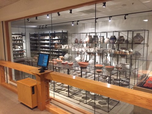 A display of various pottery pieces in a museum, viewed through glass with a wooden railing in the foreground.