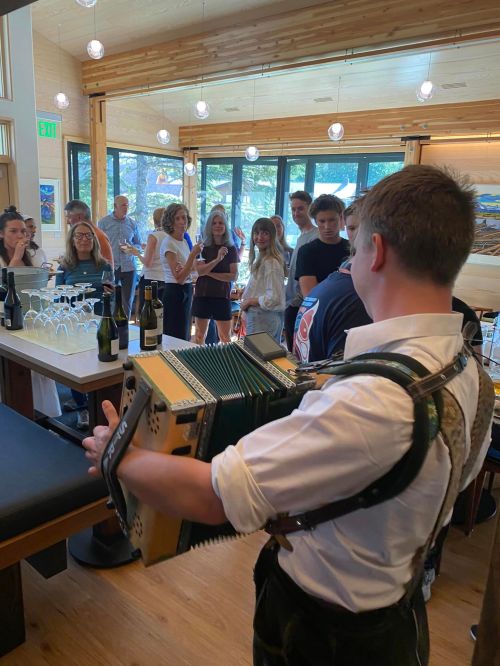 A young musician plays the accordion in a lively gathering with guests enjoying drinks in a bright, modern space.