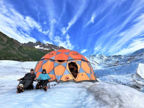 Two people relax in front of an orange and blue tent on a glacier, with mountains and a vibrant sky in the background.