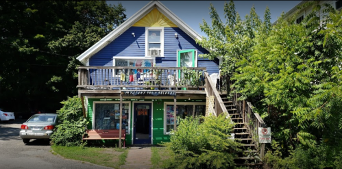 A colorful two-story building with a porch, surrounded by greenery, featuring an "Open" sign and clothes hanging outside.