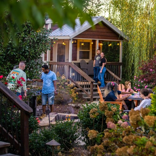 A backyard gathering with people grilling, sitting, and enjoying time near a cozy house surrounded by greenery and flowers.