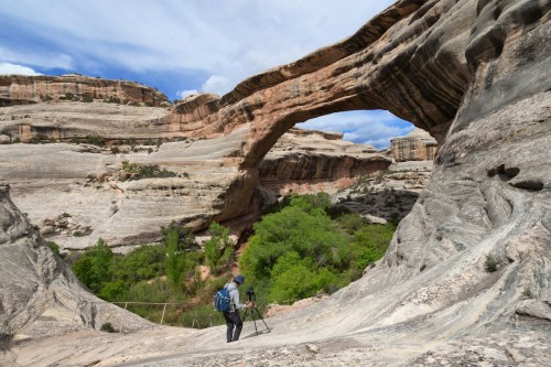 A photographer sets up a camera near a natural stone arch in a rugged landscape with greenery below.