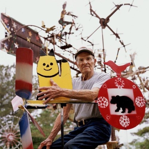 An elderly man poses with colorful metal sculptures, including a yellow cat and a bear, in a whimsical outdoor setting.