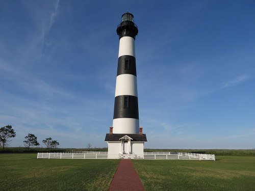 A tall black and white striped lighthouse stands against a clear blue sky, surrounded by green grass and a white fence.