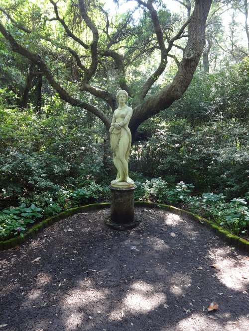 A statue of a woman stands on a pedestal surrounded by lush greenery and trees in a serene garden setting.