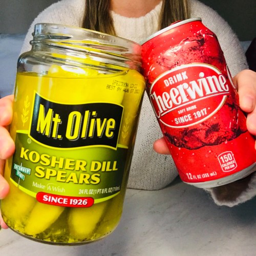 A person holding a jar of Mt. Olive Kosher Dill Spears and a can of Cheerwine soda.