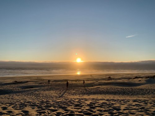 Sunset over a beach with silhouettes of people walking along the shore and gentle waves lapping at the sand.