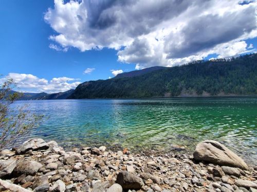 A serene lakeside view with clear blue water, rocky shore, and mountains under a partly cloudy sky.