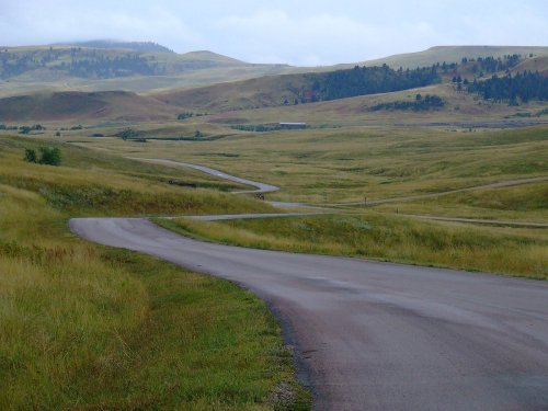 A winding road cuts through a grassy landscape with rolling hills under a cloudy sky.