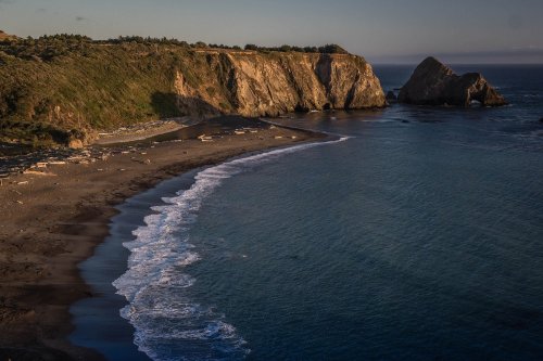 A serene beach scene with gentle waves lapping at the shore, flanked by rocky cliffs and a calm ocean at sunset.