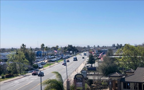 A wide street lined with trees and businesses, featuring cars and American flags under a clear blue sky.