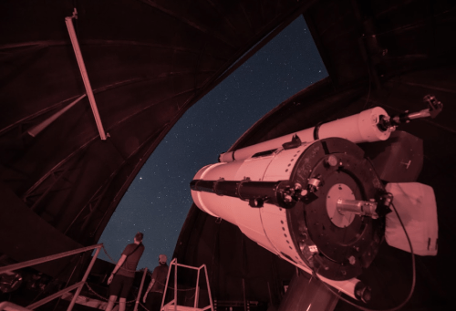 A telescope inside an observatory points towards a starry night sky, with a person observing in the background.