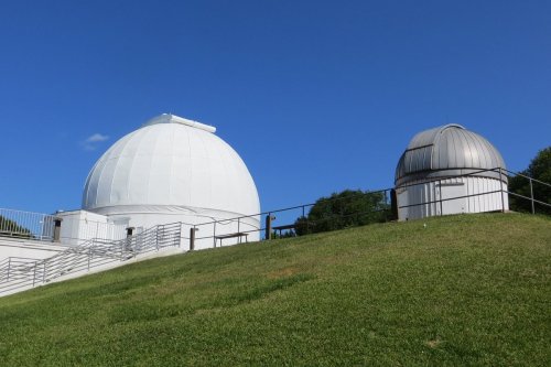 Two observatory domes on a grassy hill under a clear blue sky. One dome is large and white, the other smaller and silver.