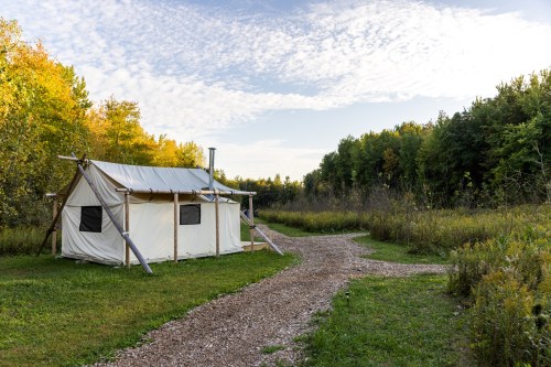 A white canvas tent sits beside a gravel path, surrounded by lush greenery and trees under a partly cloudy sky.