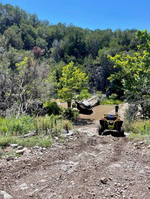 A rugged landscape with a rocky path leading to a muddy area, a person walking near a large boulder, and greenery around.
