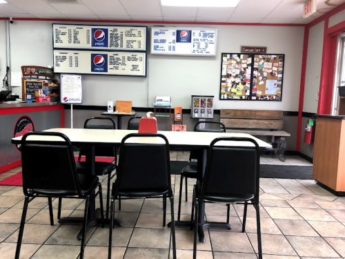 A vacant fast-food restaurant interior with a table, chairs, menu board, and bulletin board on the wall.