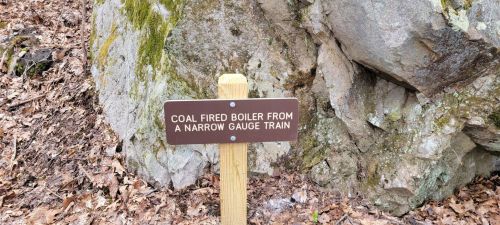 Sign on a wooden post reading "Coal Fired Boiler from a Narrow Gauge Train" near a rocky area with fallen leaves.