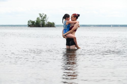 A woman holds a child in shallow water, both smiling, with a calm lake and distant trees in the background.