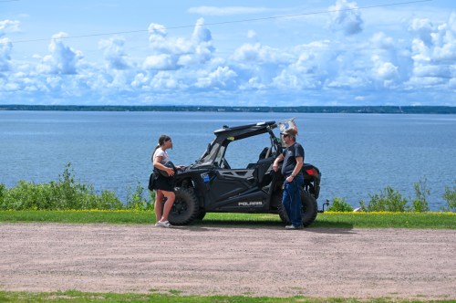 Two people stand by a black off-road vehicle near a lake, with a scenic view of clouds and greenery in the background.