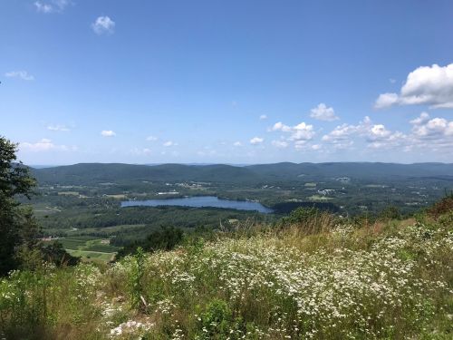 A scenic view of a lake surrounded by green hills and fields under a clear blue sky with scattered clouds.