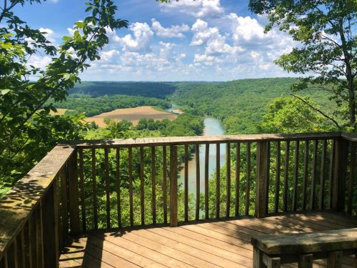 Scenic view from a wooden overlook, featuring a river winding through lush green hills under a blue sky with clouds.