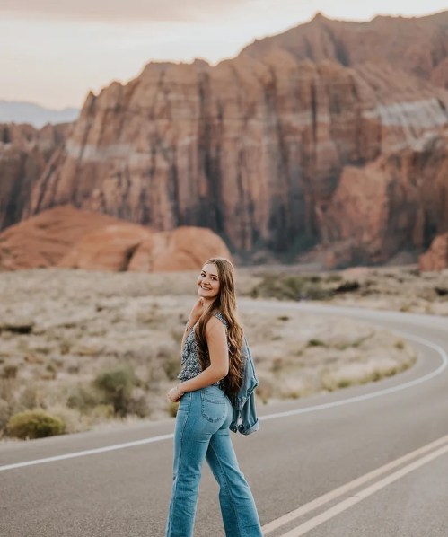 A woman with long hair stands on a winding road, smiling against a backdrop of red rock formations and mountains.