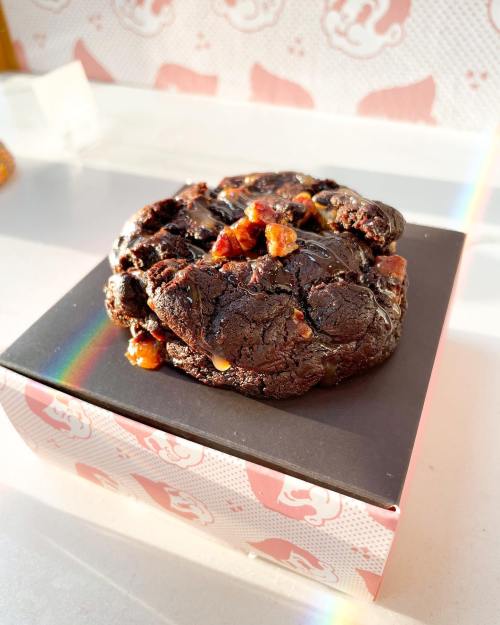 A decadent chocolate cookie topped with chunks of bacon, resting on a black box with a colorful rainbow reflection.
