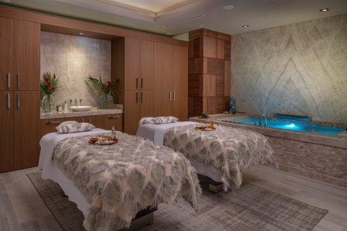 A serene spa room with two massage tables, soft linens, and a relaxing hot tub illuminated in blue light.
