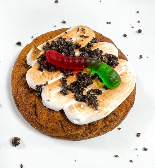 A cookie topped with marshmallow, crushed cookies, and a gummy worm, set against a white background.