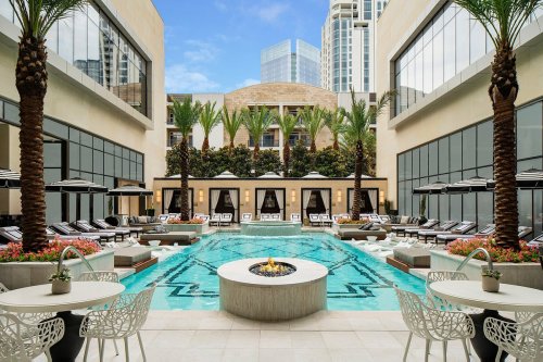 A luxurious hotel pool area with palm trees, lounge chairs, and a fire pit, surrounded by modern architecture.