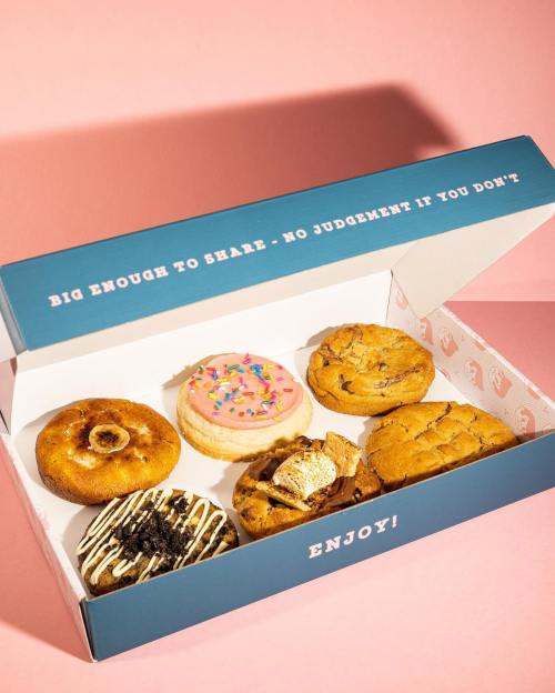 A box of assorted donuts and cookies, featuring a pink frosted donut and various cookie types, on a pink background.