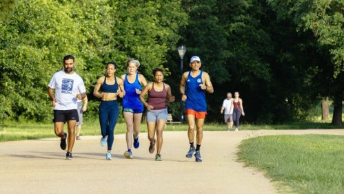 A group of five diverse runners jogs along a park path surrounded by greenery. Two people walk in the background.