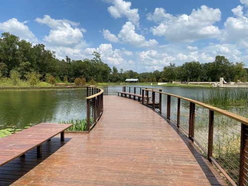 A wooden walkway curves over a calm lake, surrounded by lush greenery and a blue sky with fluffy clouds.