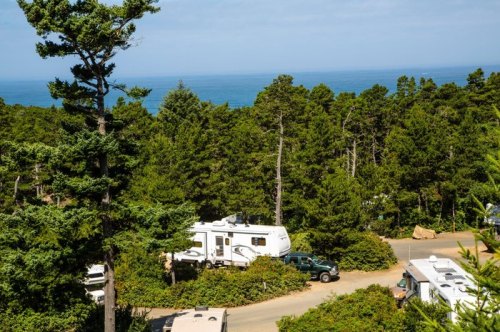 A scenic view of an RV park surrounded by trees, with the ocean visible in the background under a clear blue sky.