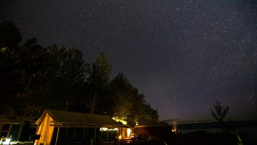 Starry night sky over a lakeside campsite, with tents and trees illuminated by soft lights.