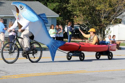 A person on a bike pulls a kayak with a person inside, while a giant fish decoration is attached to the bike.
