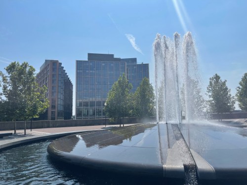 A modern building behind a large fountain, with water spraying and trees lining the walkway under a clear blue sky.