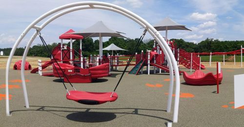 A colorful playground featuring red equipment, swings, and shaded areas under a blue sky.