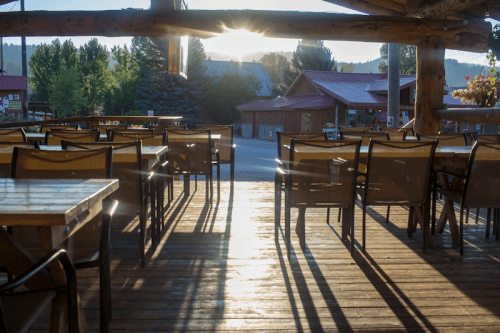 Sunlight streams through a rustic outdoor dining area, casting long shadows on wooden floors and empty chairs.