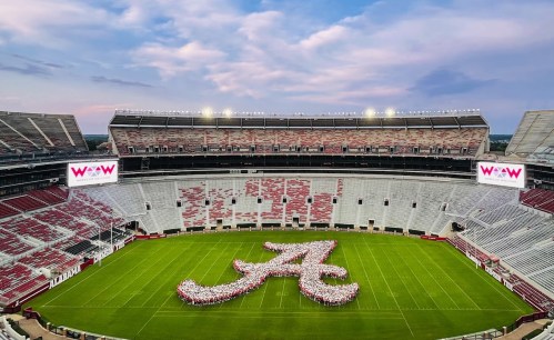Aerial view of a stadium with a large "A" formation made of people on the field, under a cloudy sky.