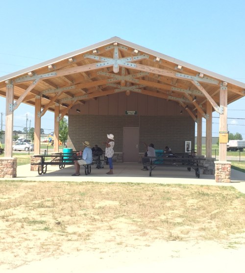 A shaded pavilion with wooden beams, featuring picnic tables and people sitting, against a clear blue sky.