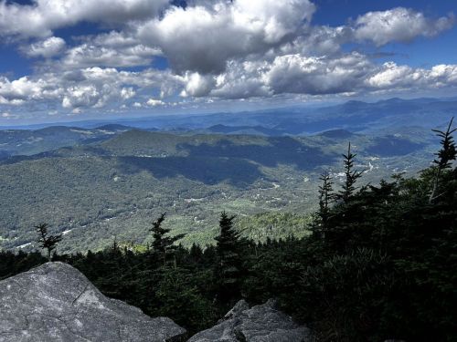 A panoramic view of lush green mountains under a cloudy blue sky, with rocky foreground and distant hills.