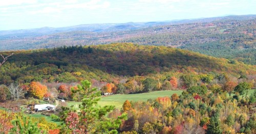 A panoramic view of rolling hills covered in vibrant autumn foliage, with a small farm nestled in the valley below.