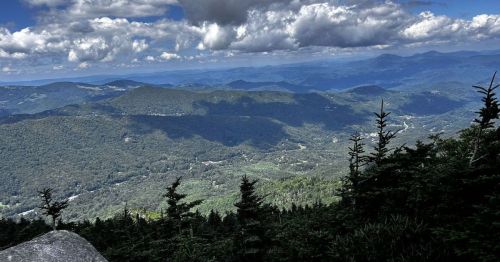 A panoramic view of lush green mountains under a partly cloudy sky, showcasing rolling hills and distant peaks.
