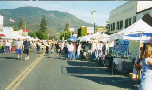 A bustling street market with tents and vendors, surrounded by mountains and a crowd of people enjoying the day.