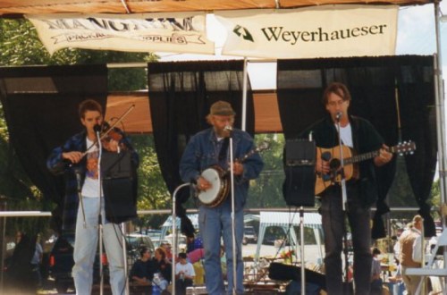Three musicians perform on stage, playing a fiddle, banjo, and guitar at an outdoor event. Banners are visible above.